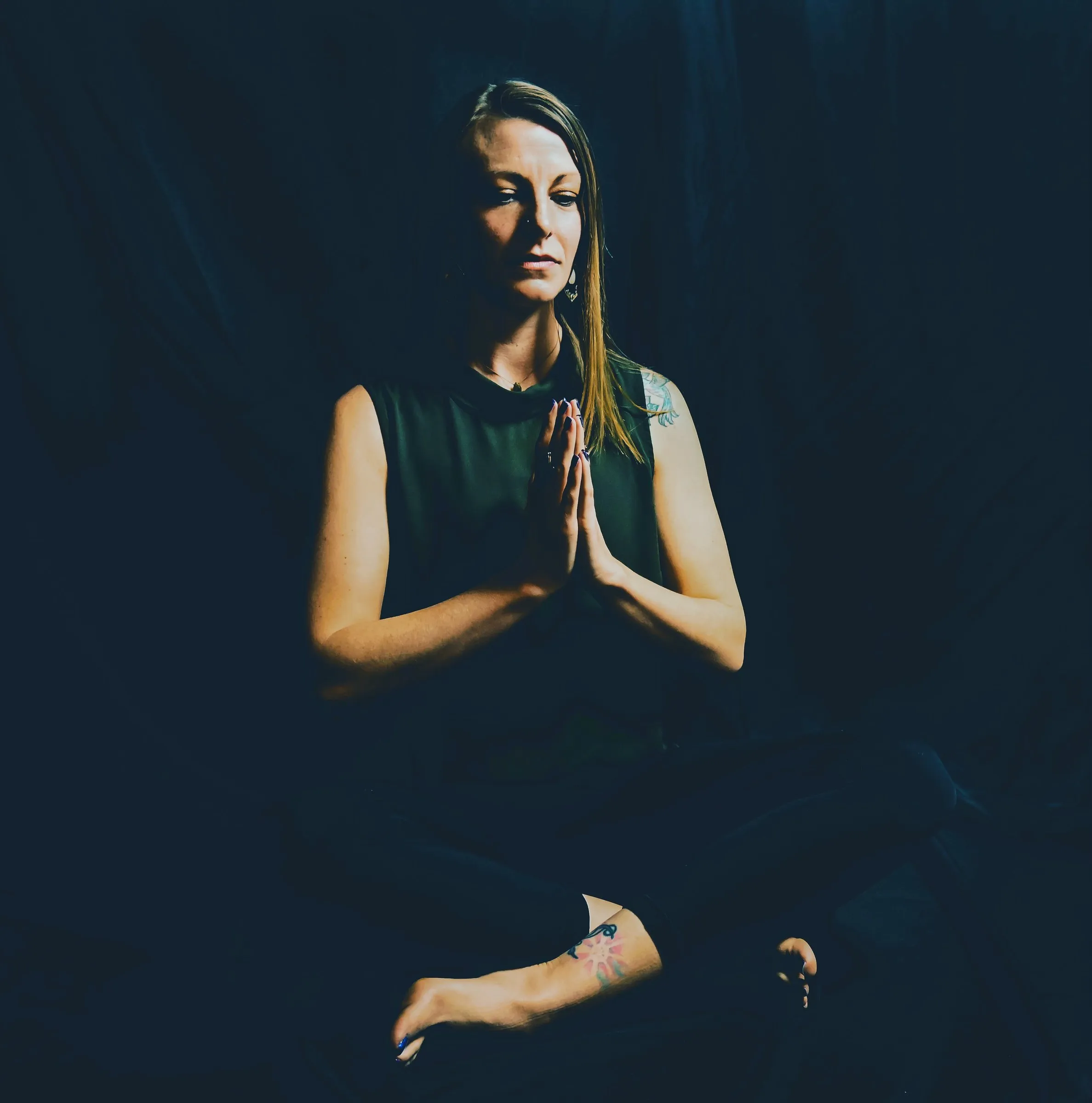 Cora seated in meditation with hands at heart center, in low dramatic light