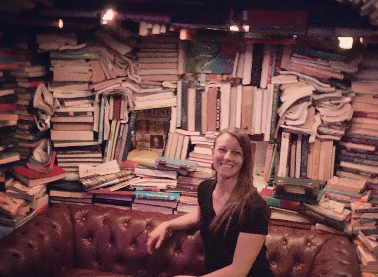Cora smiling on a couch in front of a dramatically overstuffed, disorganized wall of stacked books